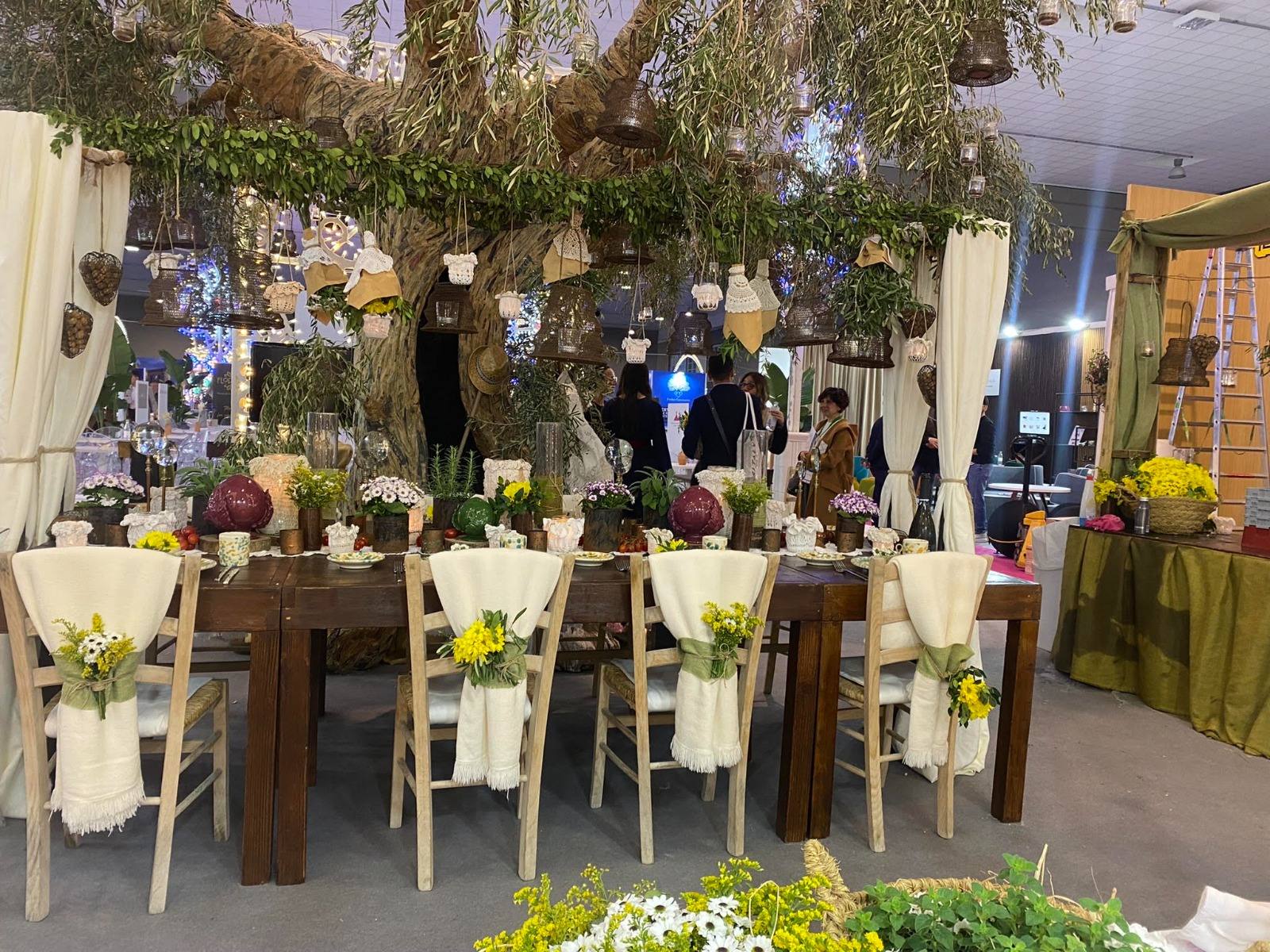 Tavola nuziale allestita sotto un ulivo, con lanterne sospese e fiori di campo